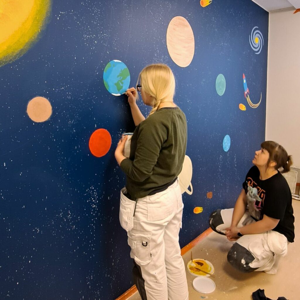 Maalariopiskelijat maalaamassa koristemaalauksia päiväkodissa.
